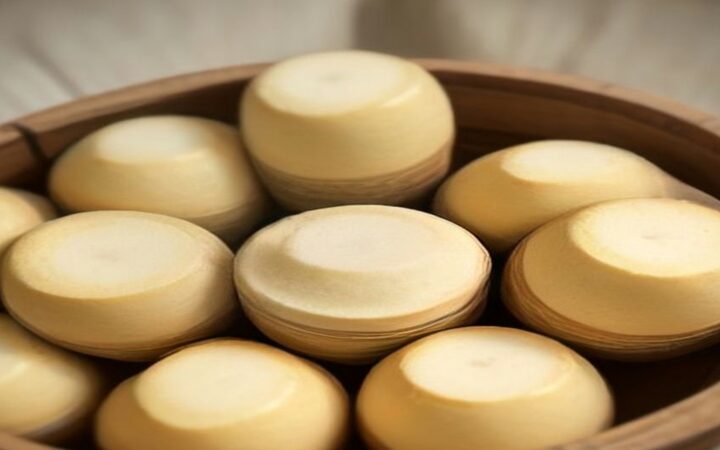 Ilustración de varias piezas redondas de queso artesanal madurado, de corteza amarillo pálido, acomodadas dentro de un recipiente de madera bajo una luz cálida y suave