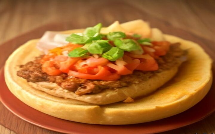 Ilustración de una torta ahogada tapatía partida a la mitad sobre un plato hondo, bañada en salsa roja de jitomate, con birote crujiente, carnitas doradas, rodajas de cebolla y limón, sobre una mesa de madera con un jarro de barro